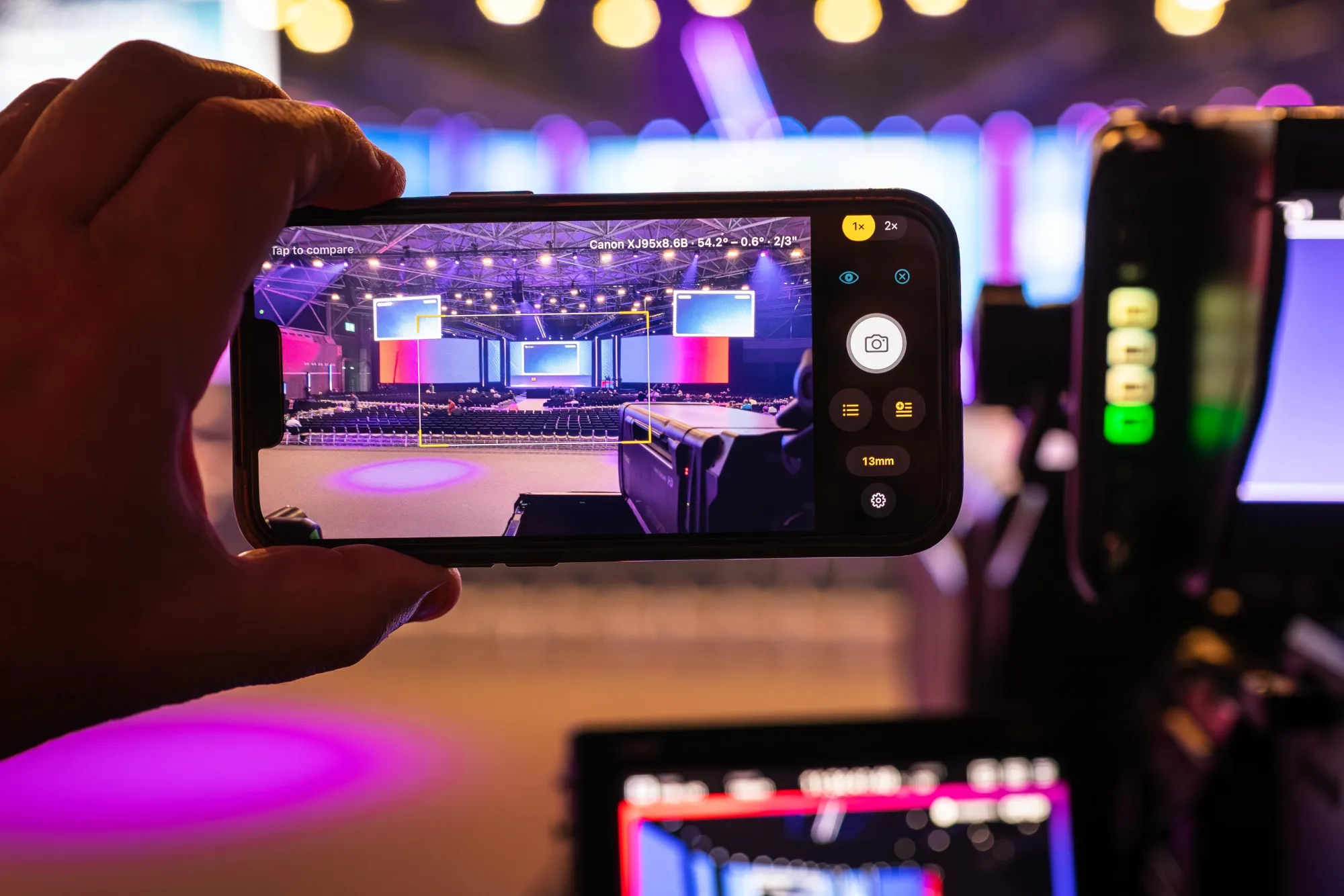 PocketOP on an iPhone held up at a live event venue, showing the wide and tele field-of-view brackets for a Canon XJ95x8.6B broadcast lens, with a broadcast camera in the foreground.
